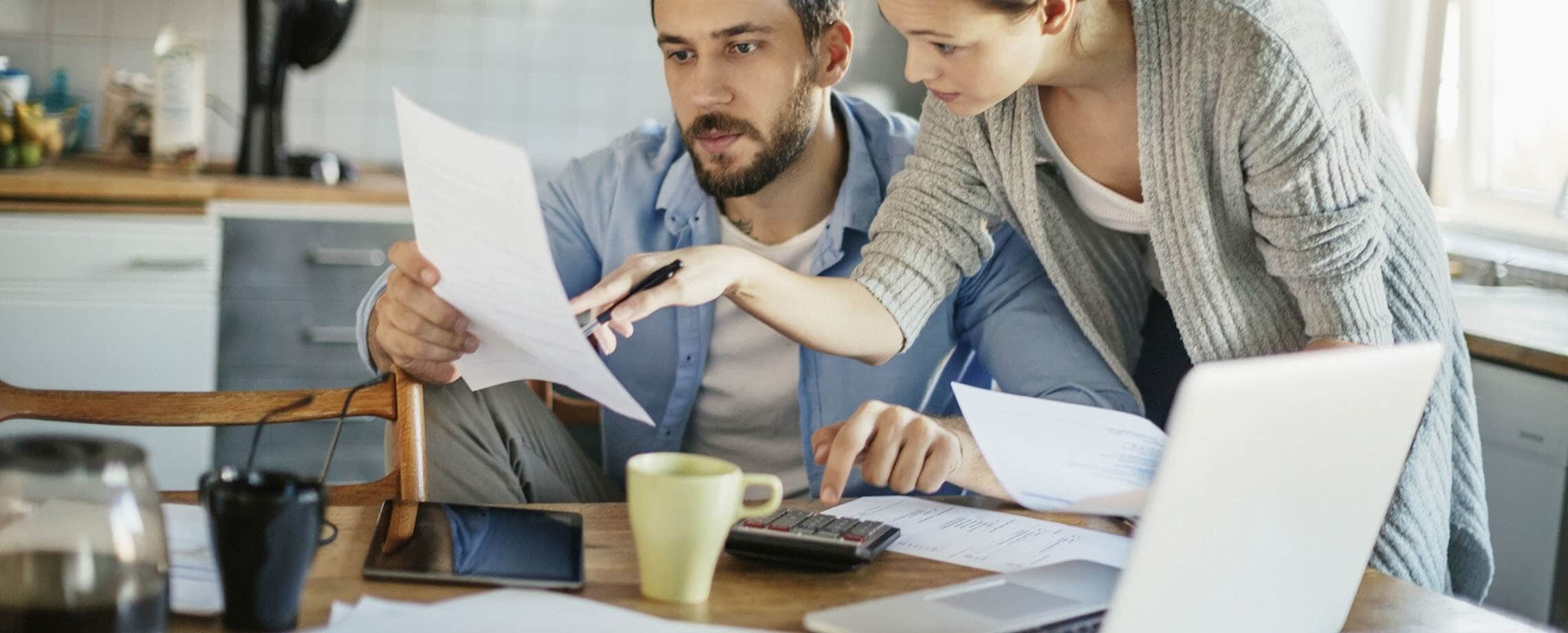 A couple discussing papers while typing numbers into a calculator at a kitchen table