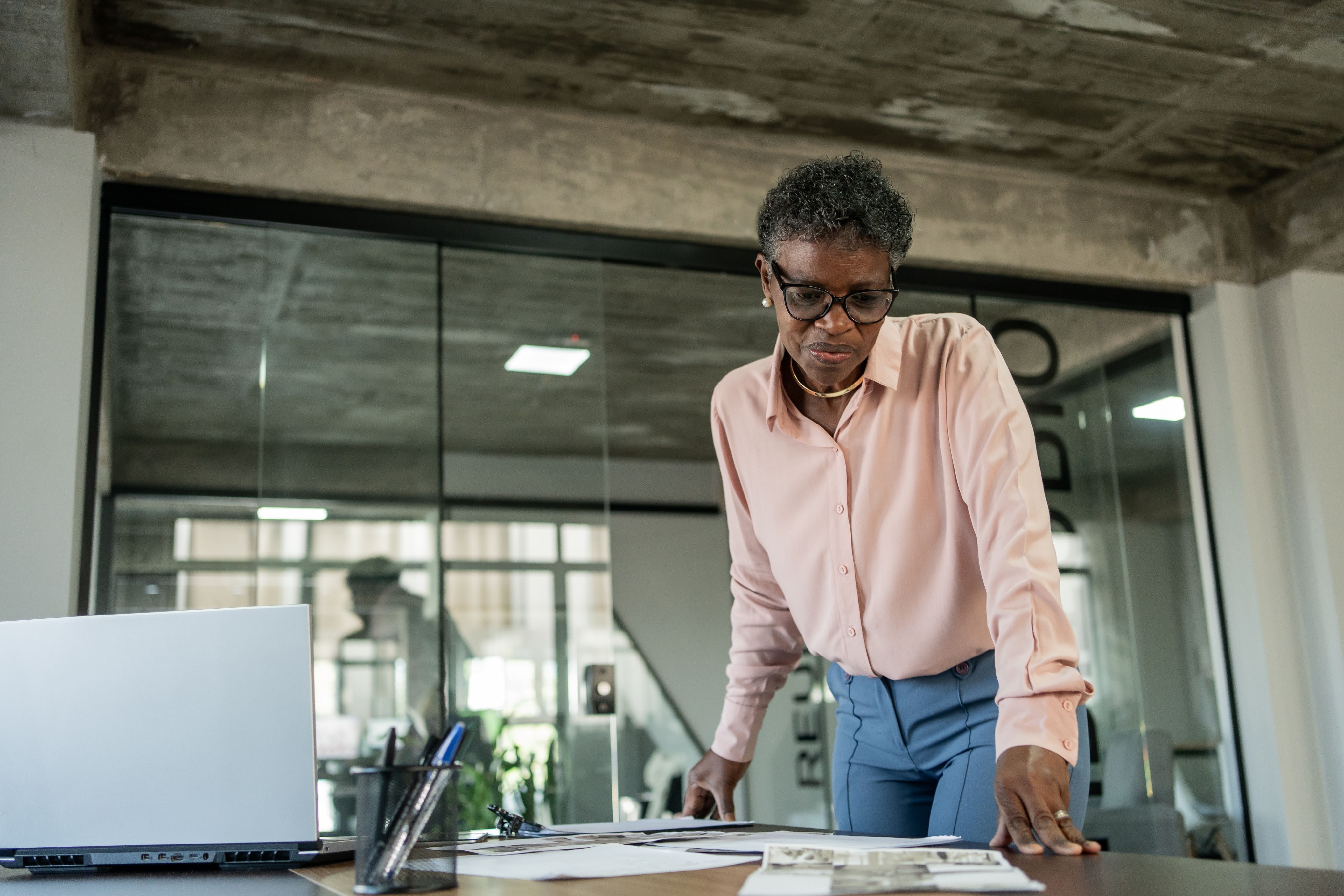 Female in pink long sleeve button up hovering over table analyzing work that is scattered across