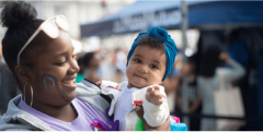 Woman wearing sunglasses on top of her head and a rainbow painted on her cheek, holding a baby and smiling