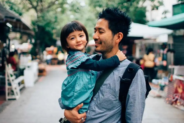 A man, holding a young girl in his arms while looking at her and smiling, walking through a city market