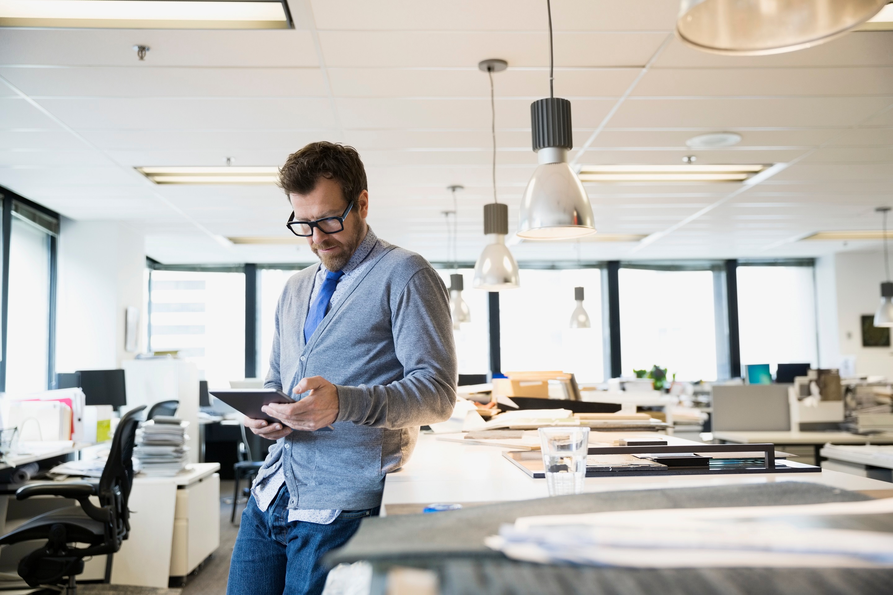 Man leaning on a desk in an open office looking at a tablet