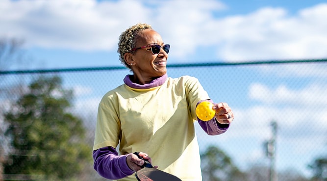 A woman, wearing a yellow shirt and sunglasses, holding a wiffle ball while standing on a tennis court outside, smiling 