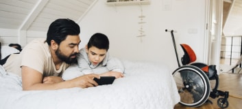 Dad with disabilities is laying on bed with his son looking at a cell phone